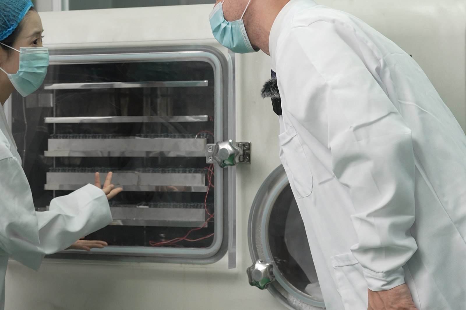 Visiting buyer in a lab coat and hairnet leaning in to inspect the shelves of a lyophilizer with a technician