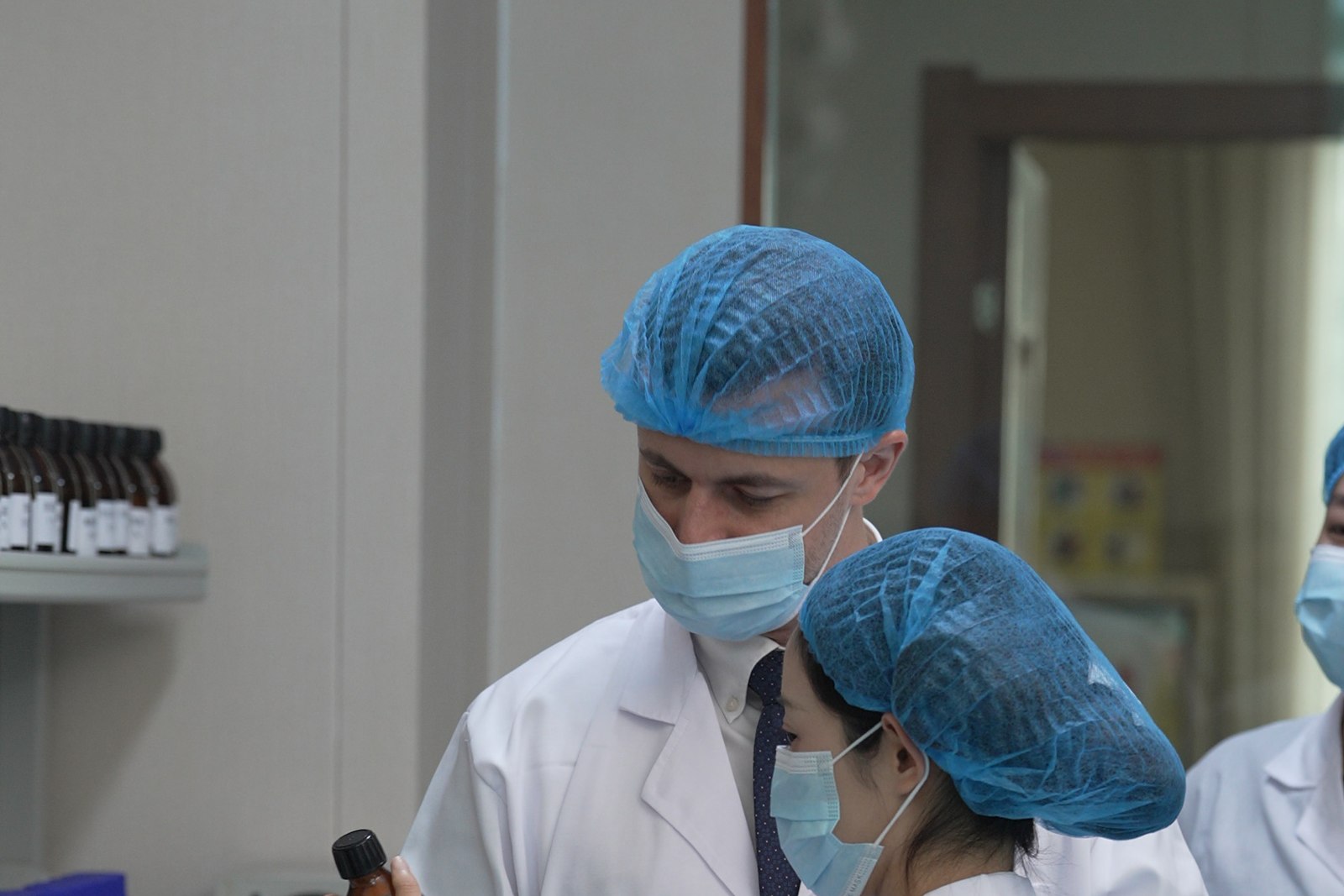 Visiting buyer and technician in hairnets and masks examining a product bottle beside an analytical balance